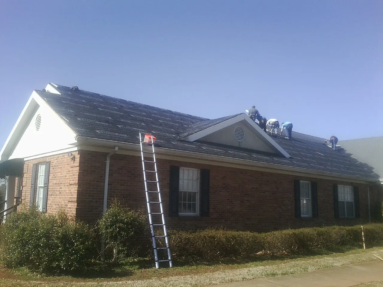 Crew tearing shingles off a brick ranch roof with a ladder set — Emergency Roof Repair work by our crew in Strongstown