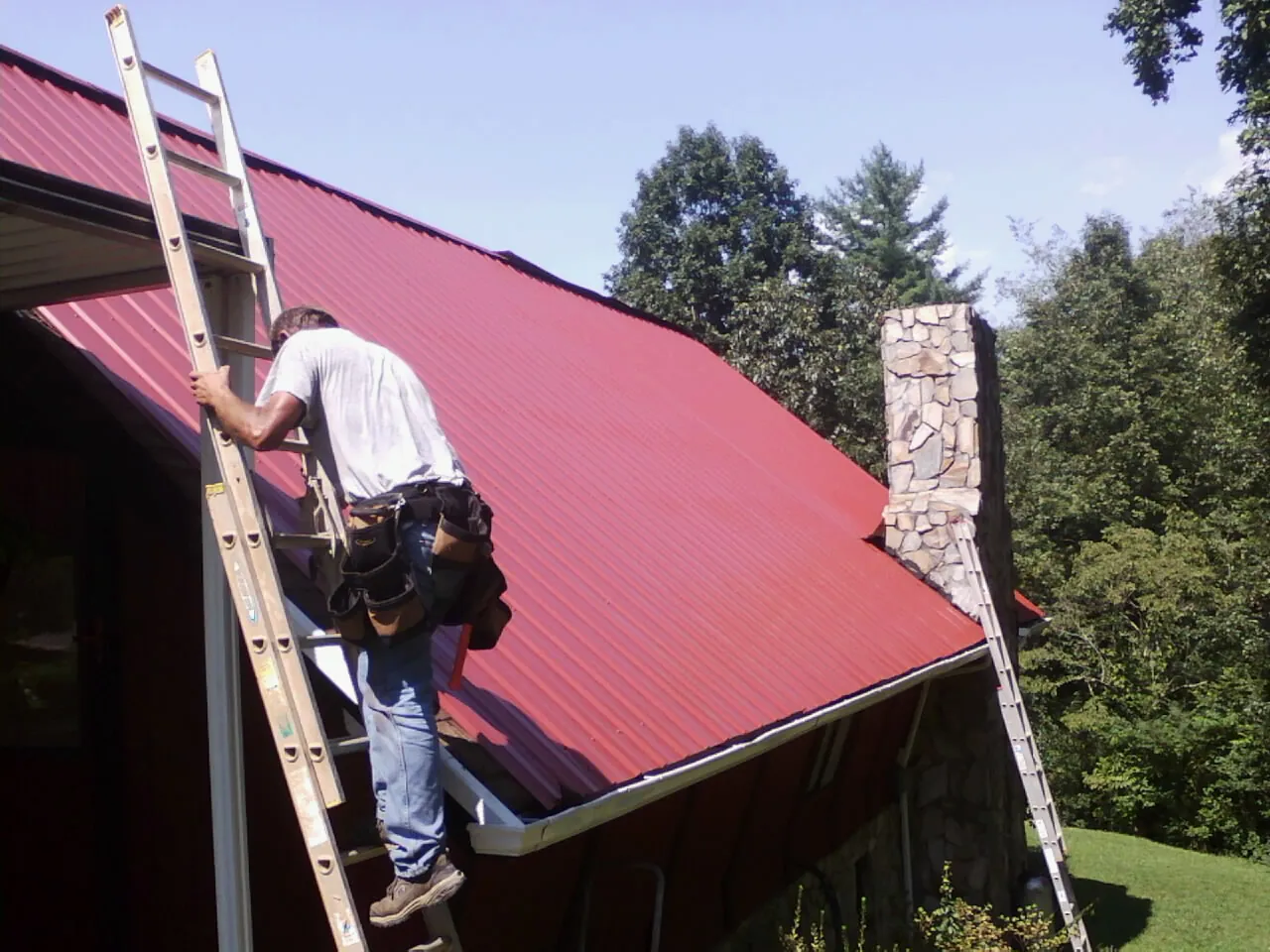 Roofer on a ladder beside a red metal roof and stone chimney — Soffit & Fascia Repair by Roofing Brothers in Strongstown